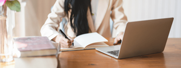 Someone sitting at a desk, writing in a notebook with a laptop open in front of them.