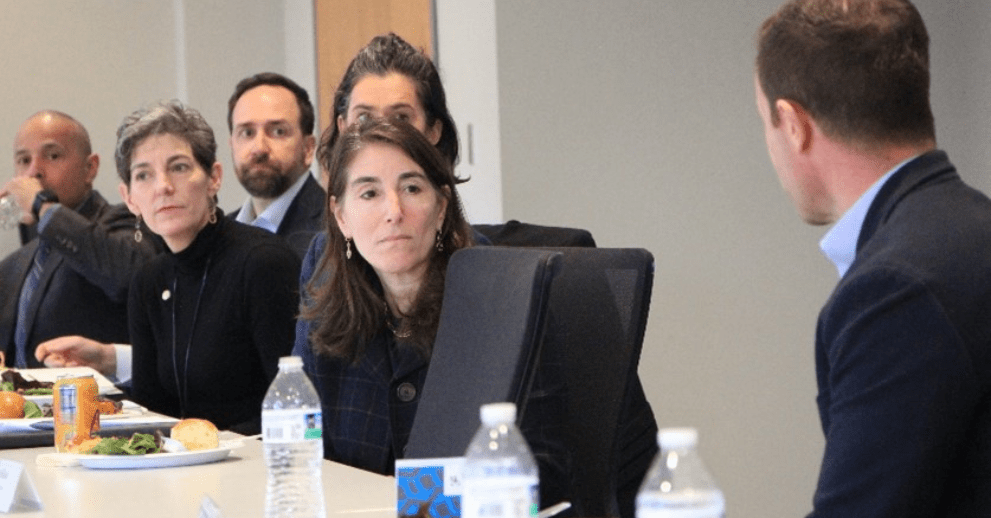 A group of people sitting at a table in a conference room listen to a man talking.