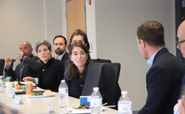 A group of people sitting at a table in a conference room listen to a man talking.