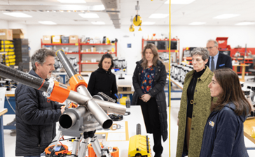 A group of people listen to a man explain a piece of equipment on a work bench in a warehouse setting.
