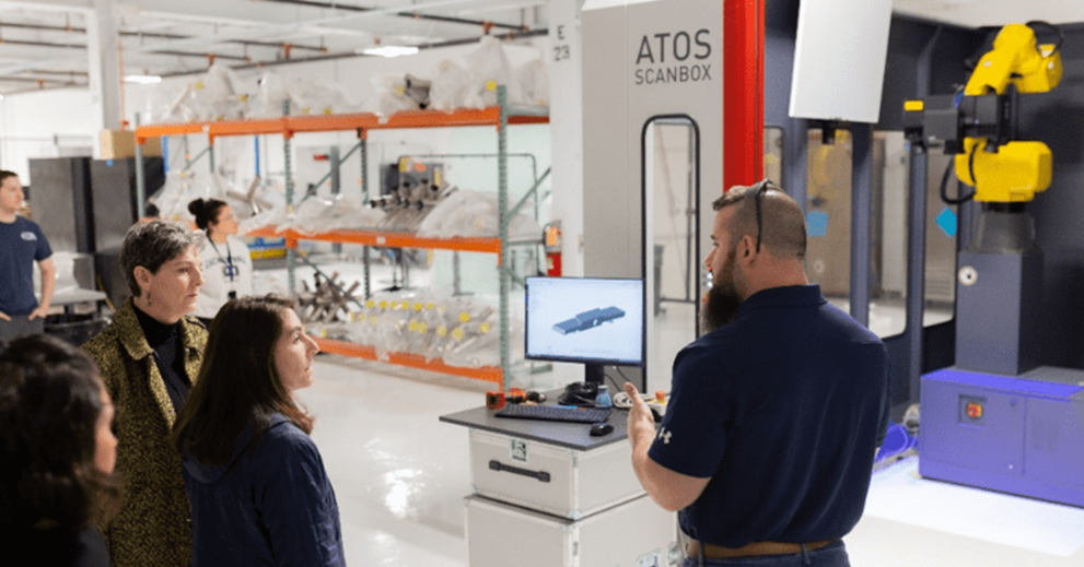 In a factory setting, a man points off in the distance as a group of people listen to him.