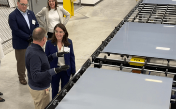 A group of people in a warehouse setting stand next to an automated manufacturing line holding large sheets of glass.
