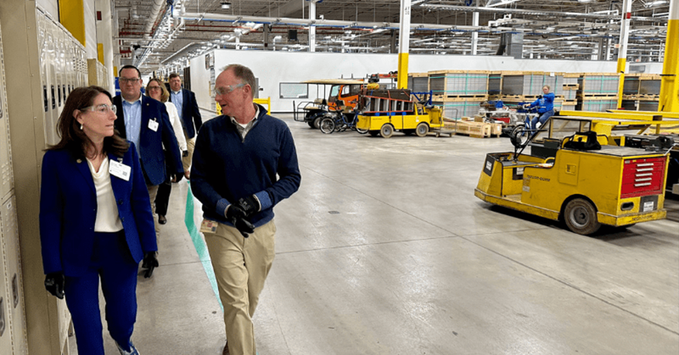 A group of people walk past large machines in a warehouse setting.