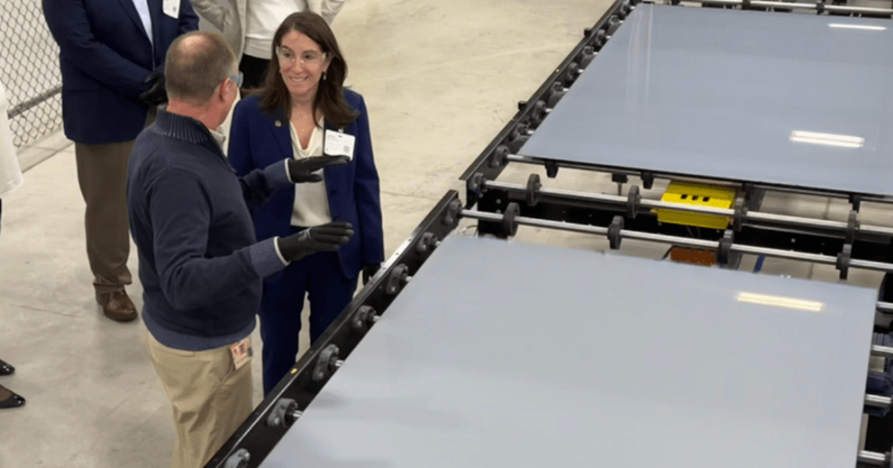 A group of people in a warehouse setting stand next to an automated manufacturing line holding large sheets of glass.