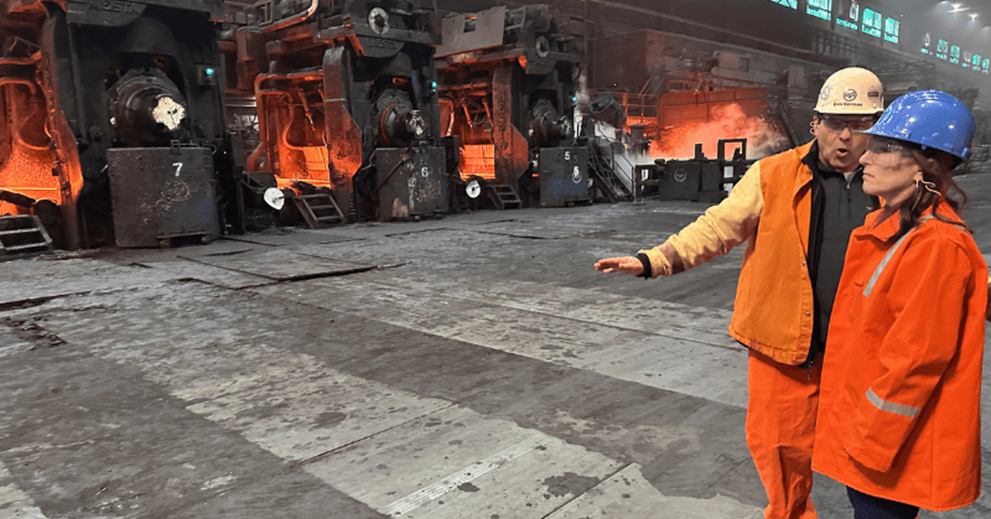 Two people in a factory setting watch a piece of molten steel