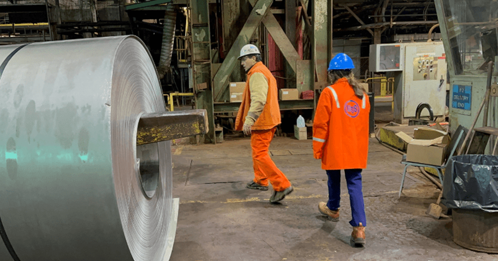 Two people in a factory setting walk past a large roll of steel
