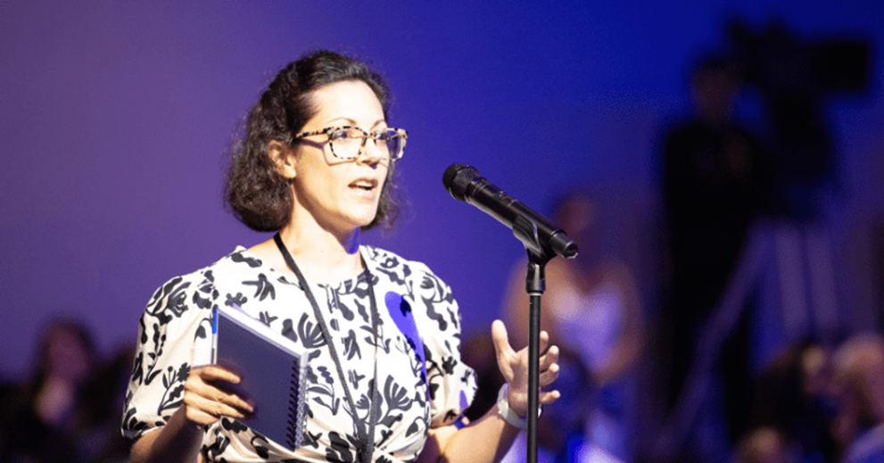 Someone talking to a room in front of a microphone with a notebook in hand.
