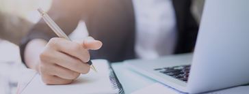 A hand holding a pen over a notebook next to a laptop on a desk.