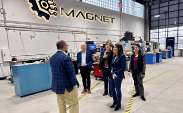 In a factory setting under a sign that says MAGNET, a group of people are on a tour.