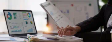 A person typing on a calculator at a desk with a laptop and papers.