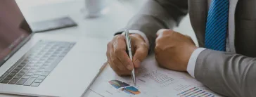 A desk with an open laptop and a person holding a pen to a paper with a pie chart.